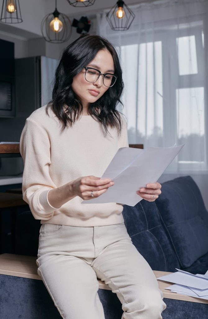 Concentrated young woman with glasses reading financial papers in a modern living room.